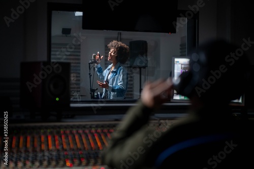 Obraz na plátně Back view of professional male sound producer with headphones is recording a new song with young african female singer in a music studio