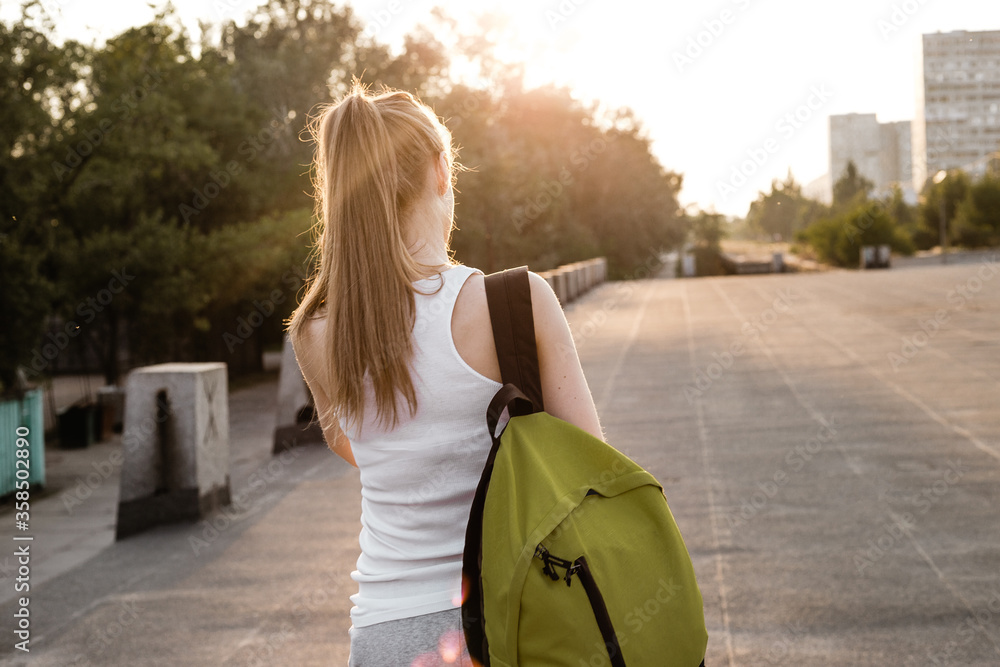 Beauty young female model with long blond hairstyle in sport clothes and with backpack standing on the road and looking sideways. The girl listen to music in earphones and walking on the cort