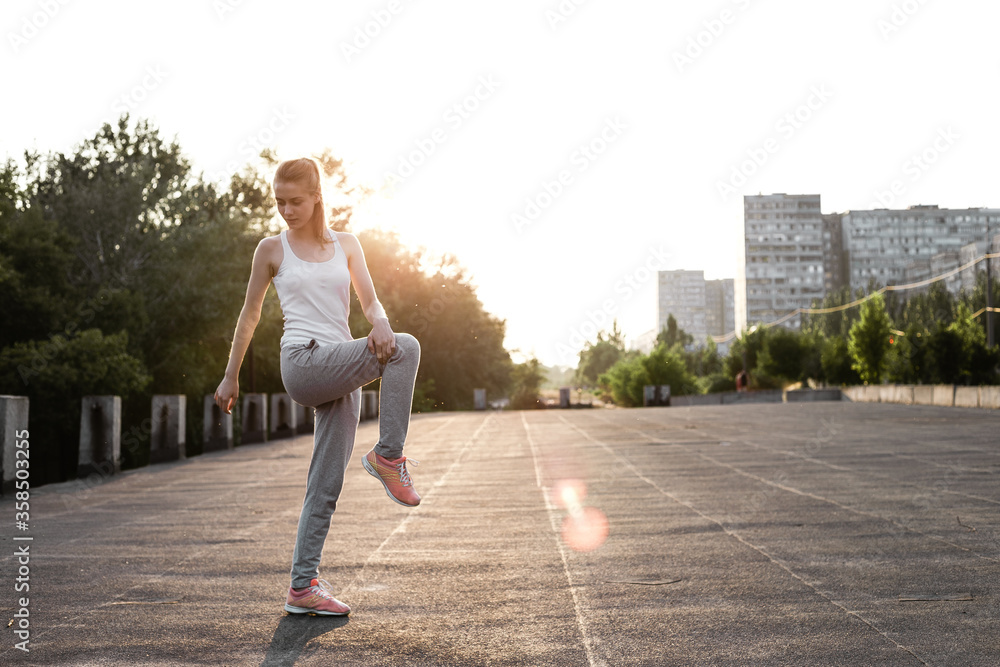 Girl stretching her legs outdoor and preparing to sport trainings. Female model posing outside on air in sporty clothes and doing exercises