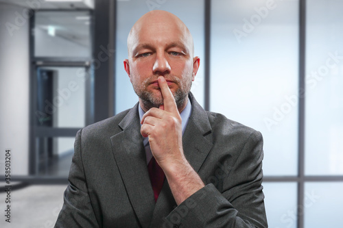Portrait of a bald unshaven businessman in grey suit on blurred office background. Hand at his face.