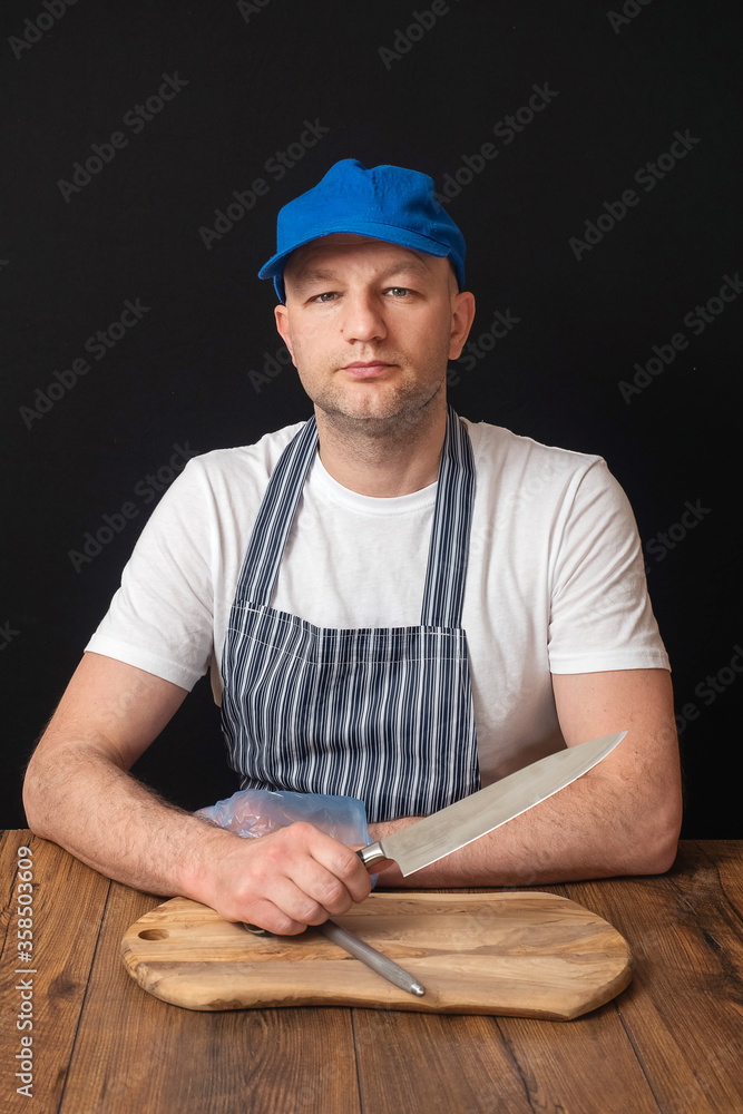 Fototapeta premium Portrait of professional butcher in white t shirt, black and white apron and blue baseball hat sitting at table and holding knife in his right hand, small cutting board in front of him.