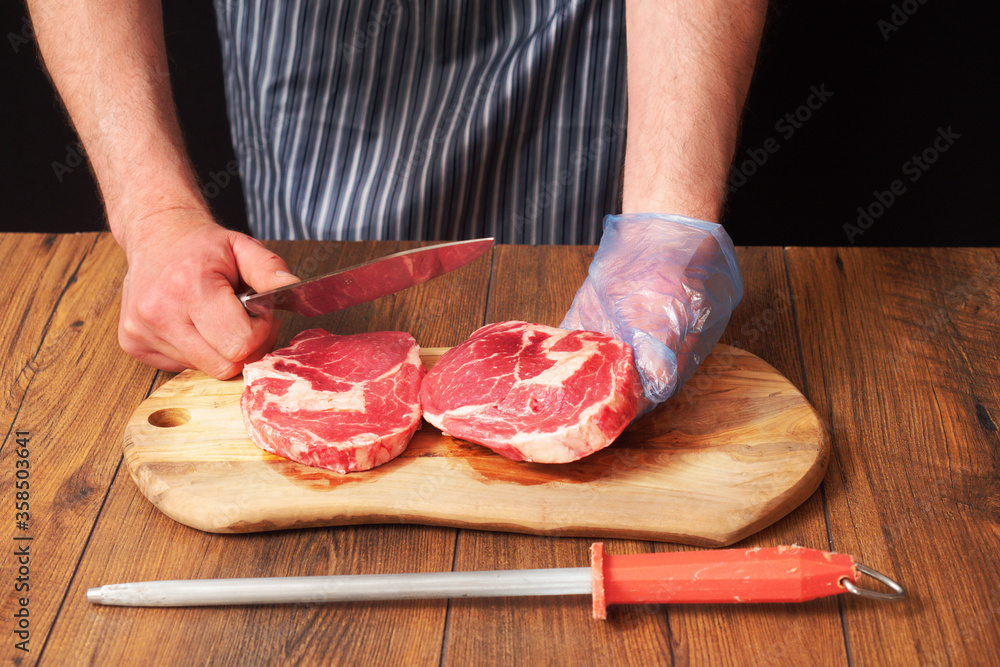 Butcher holding raw rib eye steak in at his hand, Second steak is on a wooden cutting board. Black background. Knife in butcher right hand. Steel wiht red handle in foreground.