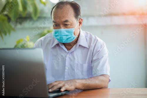 Closeup portrait of old man at home working on laptop, wearing face mask during pandemic COVID-19.