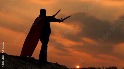 a winner with swords in his hand and in a red cloak stands on a mountain in the sunset light. free man plays superhero. game of the Roman legion. free male knight prays with outstretched swords.