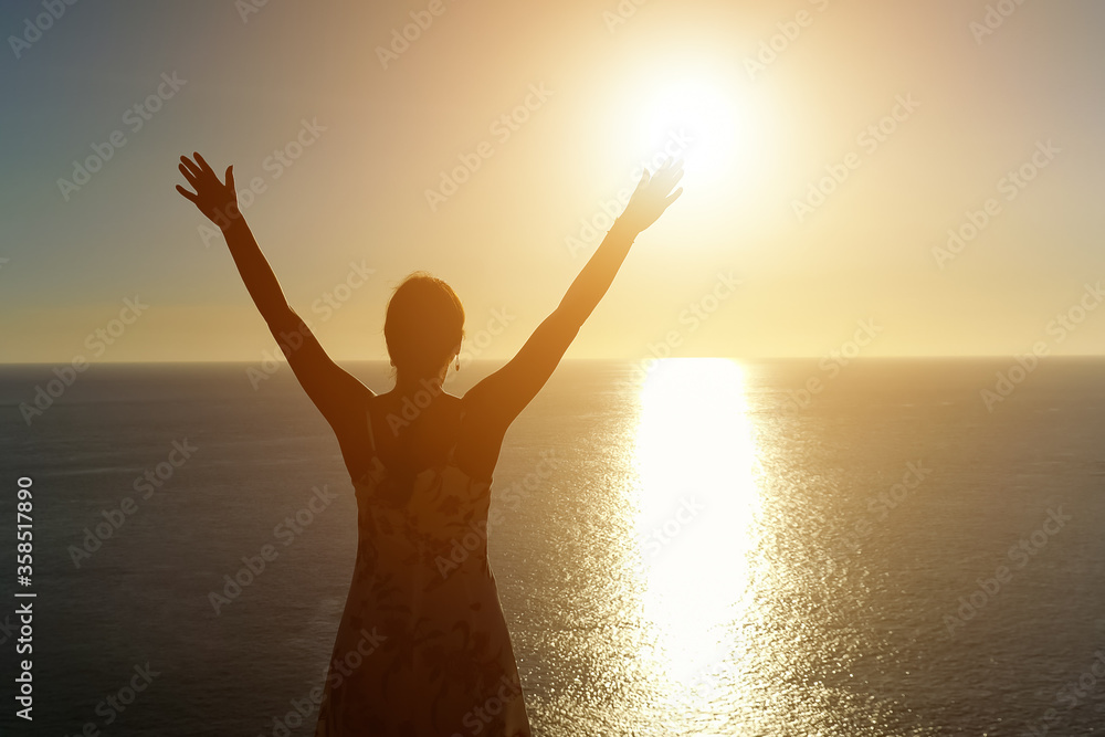 woman in summer dress silhouette stands and raises hands on ocean beach against sunset copy space