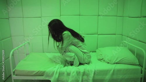 Mentally ill girl with long hair sits on a bed in a ward of a psychiatric clinic in green light. Crazy women in a straitjacket in isolation photo
