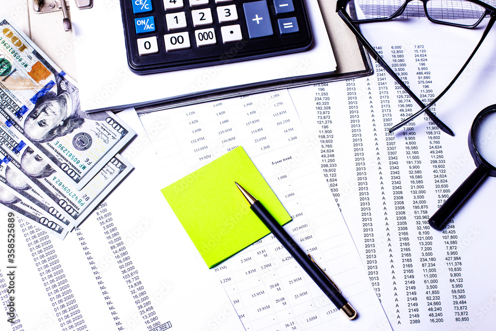Dollars, glasses, a calculator, charts, a green sticker and pen are on the desktop. Business concept top view closeup Dollars, glasses, a calculator, charts, a green sticker and pen are on the desktop. Business concept top view closeup