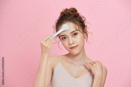Gorgeous young beautiful Asian woman applying makeup isolated over pink background.