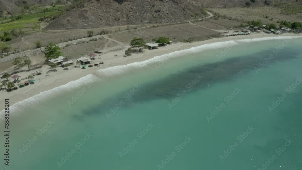 Aerial panning shot of wave splashing on shore at beach during sunny day, drone flying over turquoise sea - Lombok, Indonesia Aerial panning shot of wave splashing on shore at beach during sunny day, drone flying over turquoise sea - Lombok, Indonesia