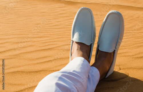 Relaxation. Man s crossed legs feet wearing white leather loafers lying in the hot desert sand.