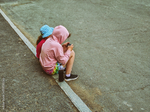 Two baby girls sitting on the side of the road and looking at a smartphone.