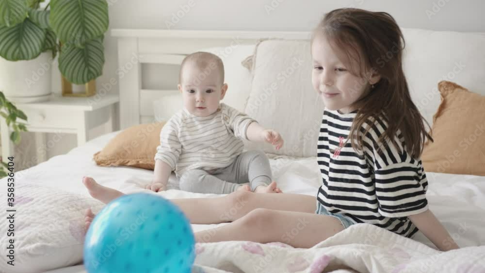 Close-Up Portrait of Two Little Sibling Baby Girls Sitting on a Bed, Playing with Blue Rubber Ball Close-Up Portrait of Two Little Sibling Baby Girls Sitting on a Bed, Playing with Blue Rubber Ball