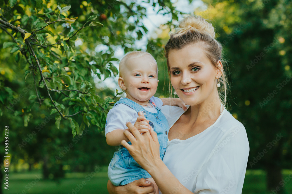 Happy smiling mother holds his little child in his arms on nature background in summer day. Portrait of young woman playing with her baby daughter. Happy motherhood. Mother's love. Happy smiling mother holds his little child in his arms on nature background in summer day. Portrait of young woman playing with her baby daughter. Happy motherhood. Mother's love.