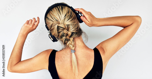 Rear view of attractive blonde young woman dancing during listening to music in headphones. Beautiful female in black dress with braid hair listens to audiobook in headphones