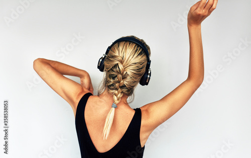Beautiful rear view of attractive blonde young woman dancing and listening to music in headphones. Joyful female in black dress with braid hair listens to favorite songs in headphones posing in studio