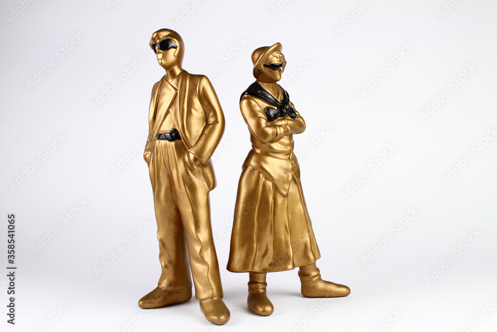 Golden plaster figures of man and woman. Vintage style. The man in a classic suit and sunglasses, the woman in a skirt and shirt with sunglasses and folded arms. White background