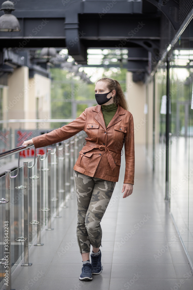 Portrait of a beautiful woman in a black mask, which stands inside a building with beautiful architecture. Woman in a brown jacket and khaki pants.