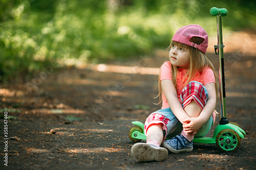 Little girl with long hair sits in a park on a scooter bending legs in knees. Close-up. Copy space