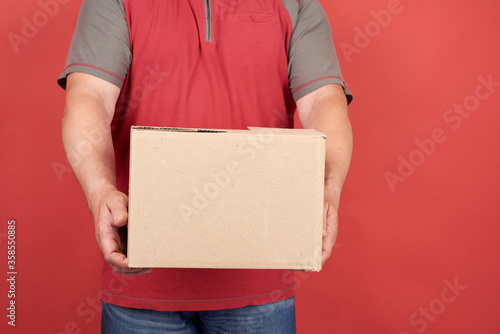 adult man in a red T-shirt holds a cardboard brown box on a red background, concept of delivery of goods