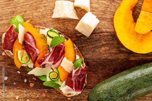 Open faced sandwich with iberico ham, parmesan and goat cheese, zucchini, pumpkin, basil and chives on sourdough bread. All on rustic chopping board, aside parmesan, pink salt, zucchini and pumpkin.