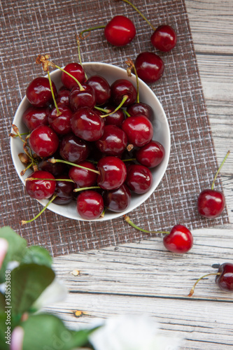 cherry berries in a white Cup, light kitchen napkin, light wooden background, blurred leaves and flowers in the foreground, flat fiy
