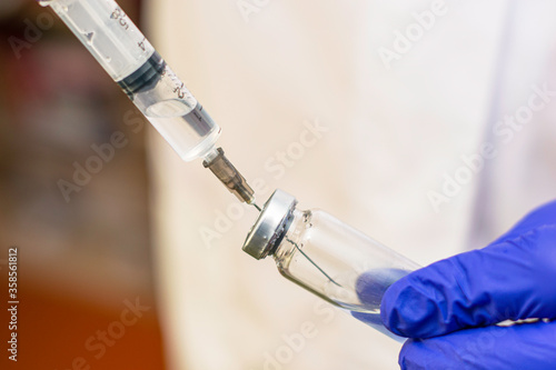 hands in blue medical surgical gloves pick up the vaccine from the ampoule with a syringe. doctor in a background in blur