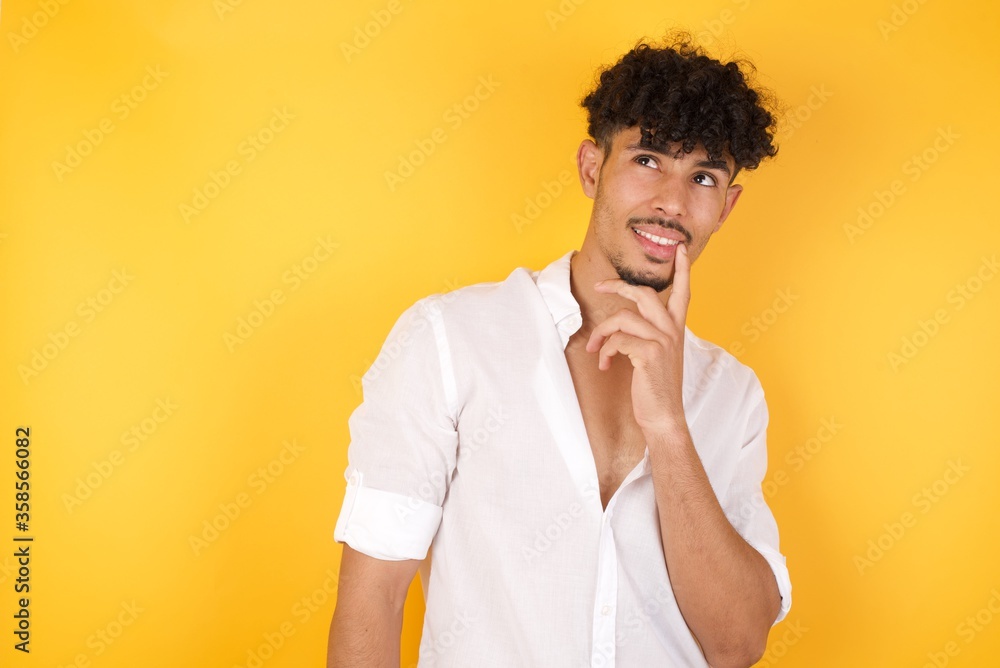 Dreamy male with thoughtful expression, looks to the camera, keeps hand near face, bitting a finger thinks about something pleasant, poses against gray wall. Dreamy male with thoughtful expression, looks to the camera, keeps hand near face, bitting a finger thinks about something pleasant, poses against gray wall.