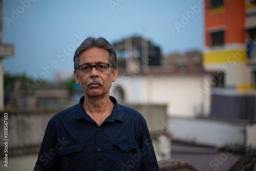 Close up portrait of an An old/aged Indian Bengali man in blue shirt is standing on a rooftop under the open sky in front of a urban landscape. Indian lifestyle and seniors