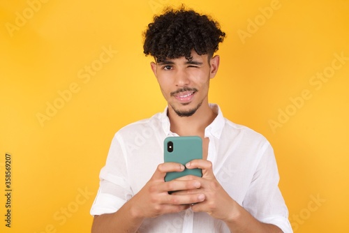 Obraz na plátně Pleased African American man wearing casual clothes using self phone and looking and winking at the camera over yellow background