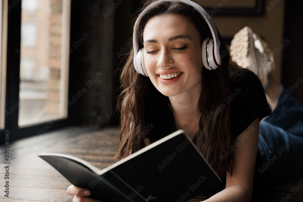 Photo of young woman wearing headphones lying on wooden floor with book Photo of young woman wearing headphones lying on wooden floor with book