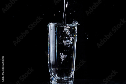 A stream of water pours into a glass of water. Water overflows the glass. Photo on a black background. Pure and purified water pouring out of a glass