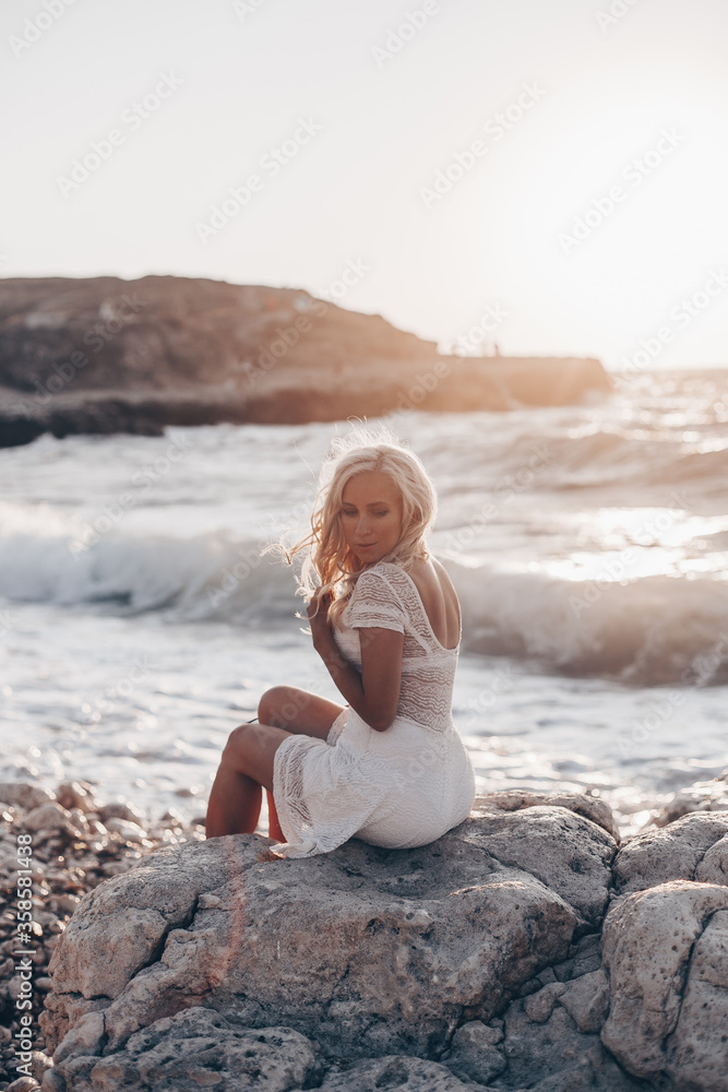 Lonely beautiful romantic girl with blonde wavy hair in white sexy dress looking at the sunset and storm on the sea and dreaming. Freedom concept