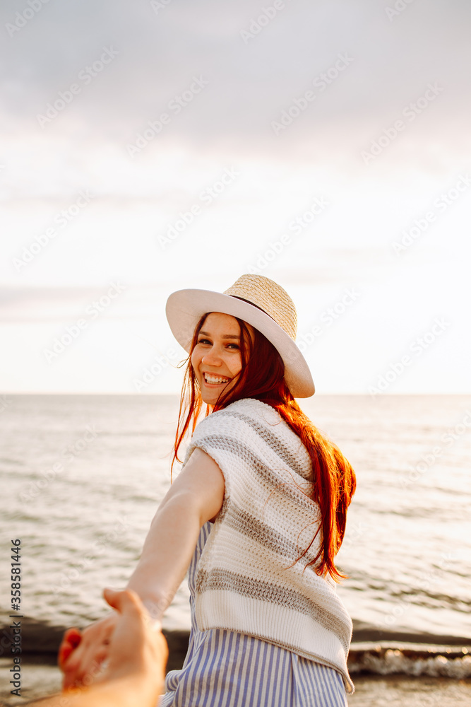 Smiling long haired woman in a hat runs holding boyfriend hand along empty ocean beach sand at sunset . Follow me concept