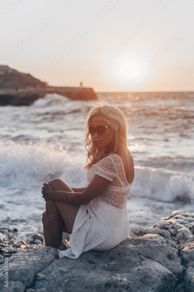 Lonely beautiful romantic girl with blonde wavy hair in white sexy dress looking at the sunset and storm on the sea and dreaming. Freedom concept
