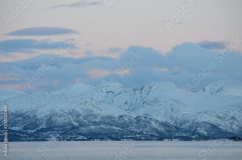 majestic snowy mountain with colourful sunset sky and beautiful cloud formation with cold fjord water underneath in wintertime