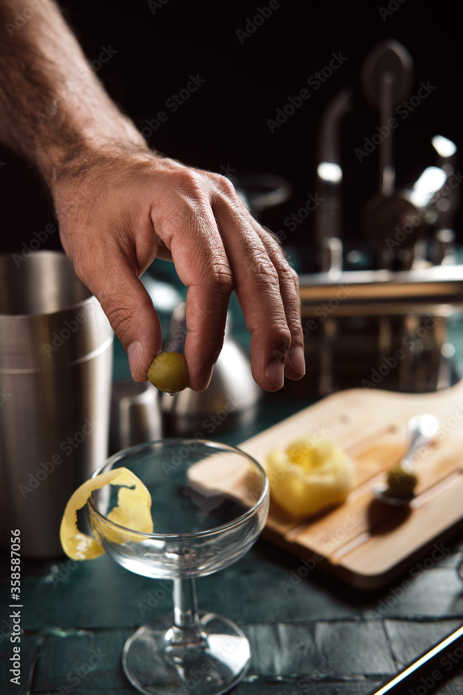 Crop anonymous male bartender putting green olive into coupe glass goblet garnished with lemon zest while preparing cocktail at bar counter Crop anonymous male bartender putting green olive into coupe glass goblet garnished with lemon zest while preparing cocktail at bar counter