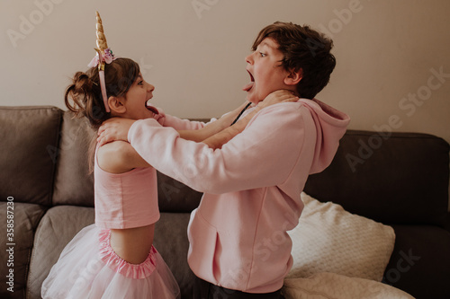 Side view of brother and sister in pink outfits strangle each other while fighting near sofa at home photo