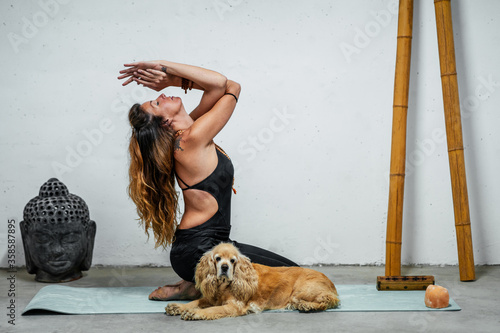 Side view of content female sitting on yoga mat with English Cocker Spaniel dog and meditating in Padmasana in room with Buddha head and bamboo sticks photo