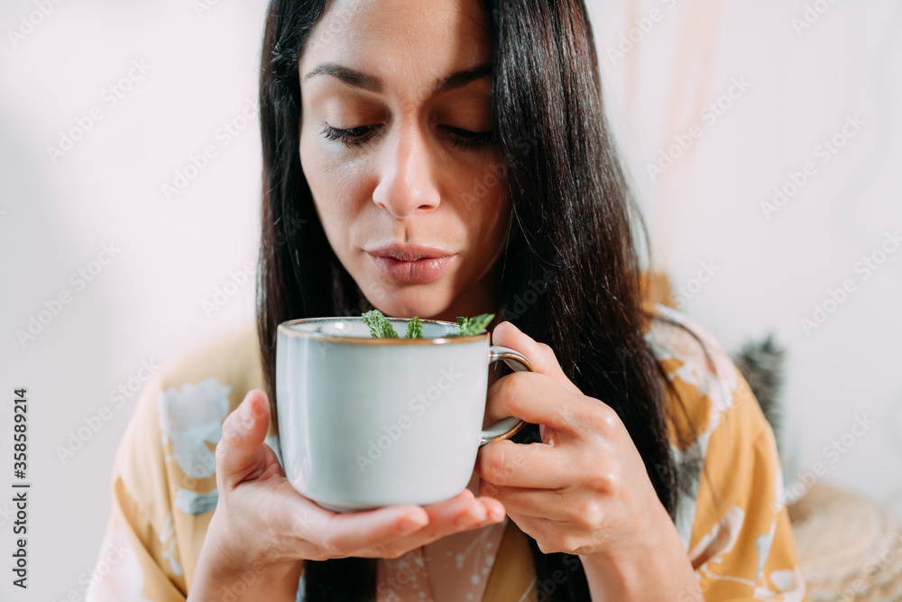 Calm relaxed brunette woman in home clothes holding mug of hot tasty tea and blowing before drinking while enjoying free time at home Calm relaxed brunette woman in home clothes holding mug of hot tasty tea and blowing before drinking while enjoying free time at home