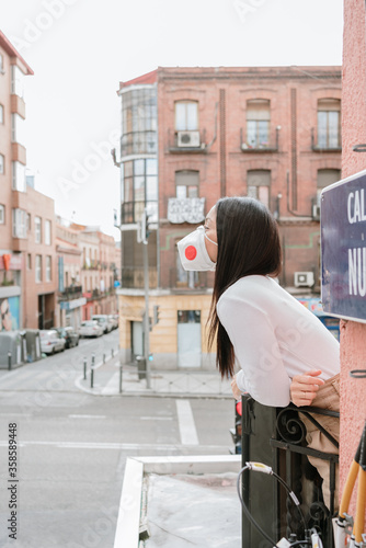 Side view of young female in casual outfit and protective mask standing on balcony in city and looking away while spending day in self isolation at home during coronavirus pandemic photo