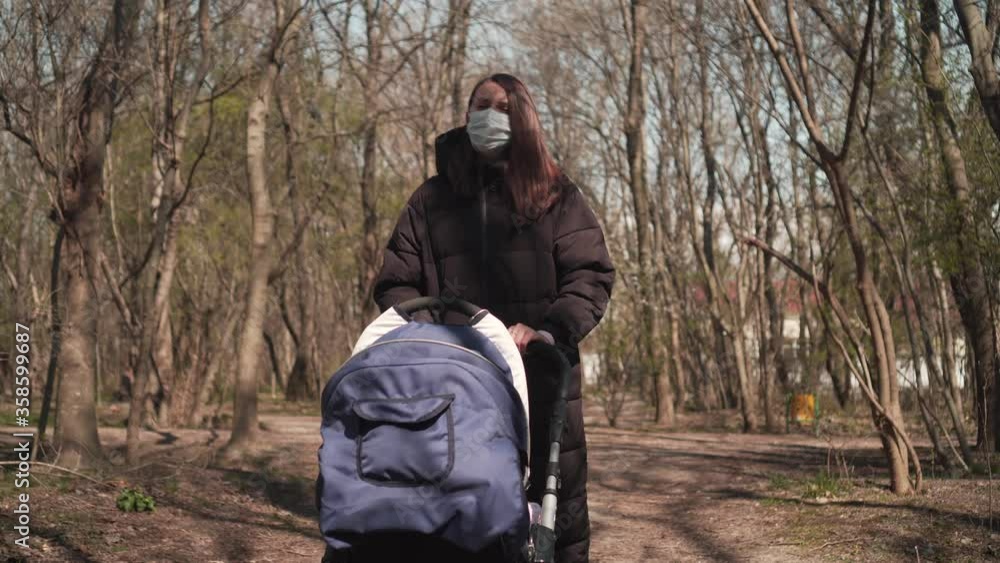 Mom in a protective mask with a pram is walking in the park among the trees. Dramatic Shot, Pandemic Virus, 4K. Mom in a protective mask with a pram is walking in the park among the trees. Dramatic Shot, Pandemic Virus, 4K.