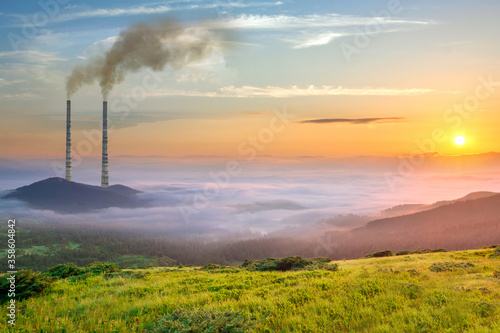 High plant pipes with thick dirty smoke emerging from wide valley with white fog between green mountains.