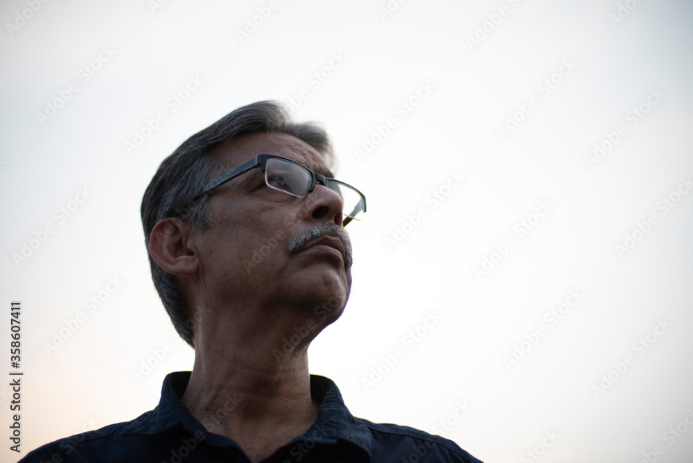 Low angle close up portrait of an An old/aged Indian Bengali man in blue shirt standing on a rooftop under the open sky copy space white background. copy space portrait Low angle close up portrait of an An old/aged Indian Bengali man in blue shirt standing on a rooftop under the open sky copy space white background. copy space portrait