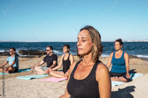 Group of several people practicing yoga and meditation on a beach in sunny day. The group is made up of several men and women of different ages.