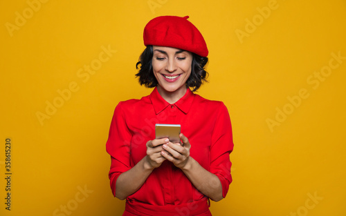 Nice photo! Half-length photo of a pleased french style dressed woman in a red beret, being in a vojage, looking at her selfie in a phone. photo