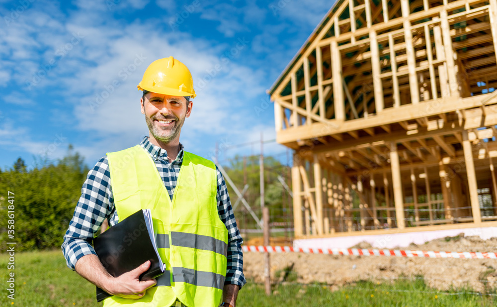Engineer with hardhat and blueprints on building site of wood frame house under construction Engineer with hardhat and blueprints on building site of wood frame house under construction