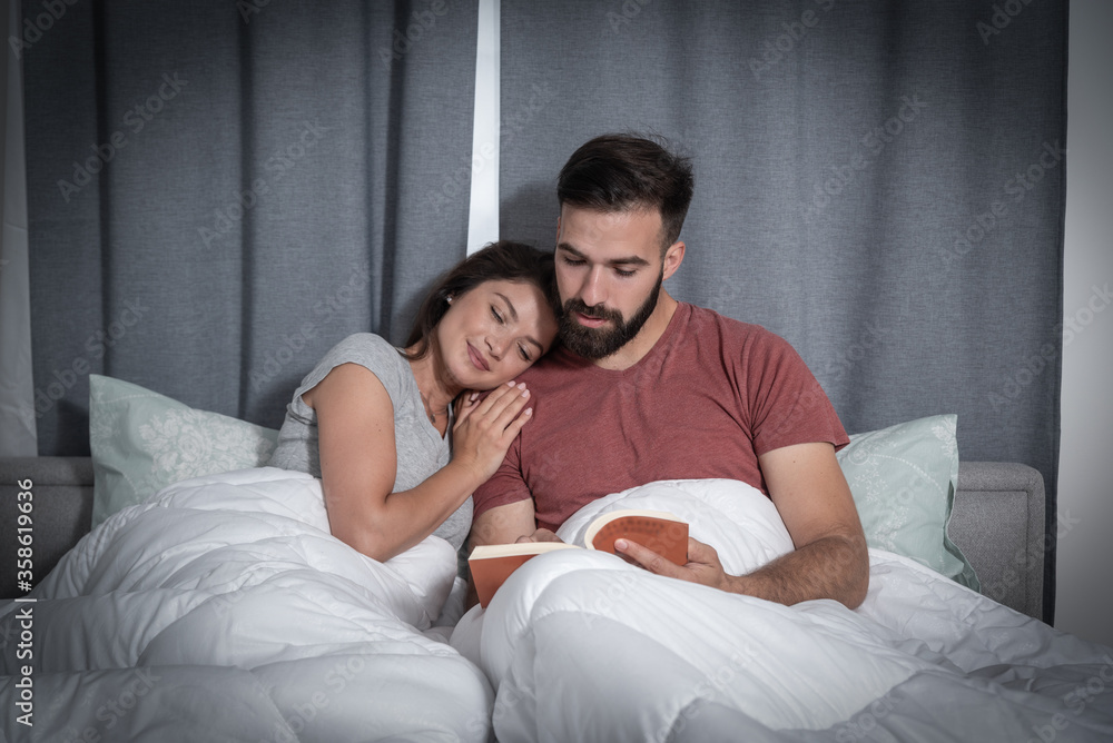 Young couple man and woman lying in bed he is reading a book to her while she leaned her head on his shoulder and enjoyed it Young couple man and woman lying in bed he is reading a book to her while she leaned her head on his shoulder and enjoyed it
