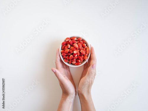 Fresh ripe red wild strawberries in a white ceramic bowl and woman hands on a white background. Top view