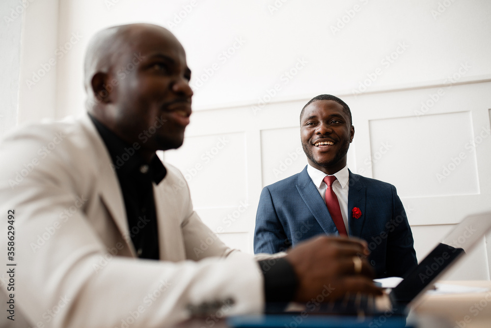 Portraits of two black men in suits sitting next to each other at the office at the table and talking to each other. Portraits of two black men in suits sitting next to each other at the office at the table and talking to each other.