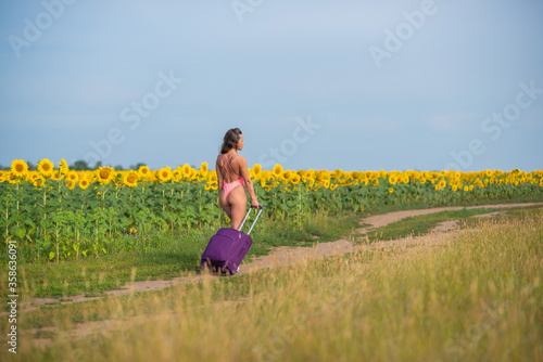 Beautiful woman in a pink swimsuit travels through the countryside with a suitcase. A girl in a monokini is walking alone on a field of sunflowers with a big bag. Hiking. Rest and social distance. photo